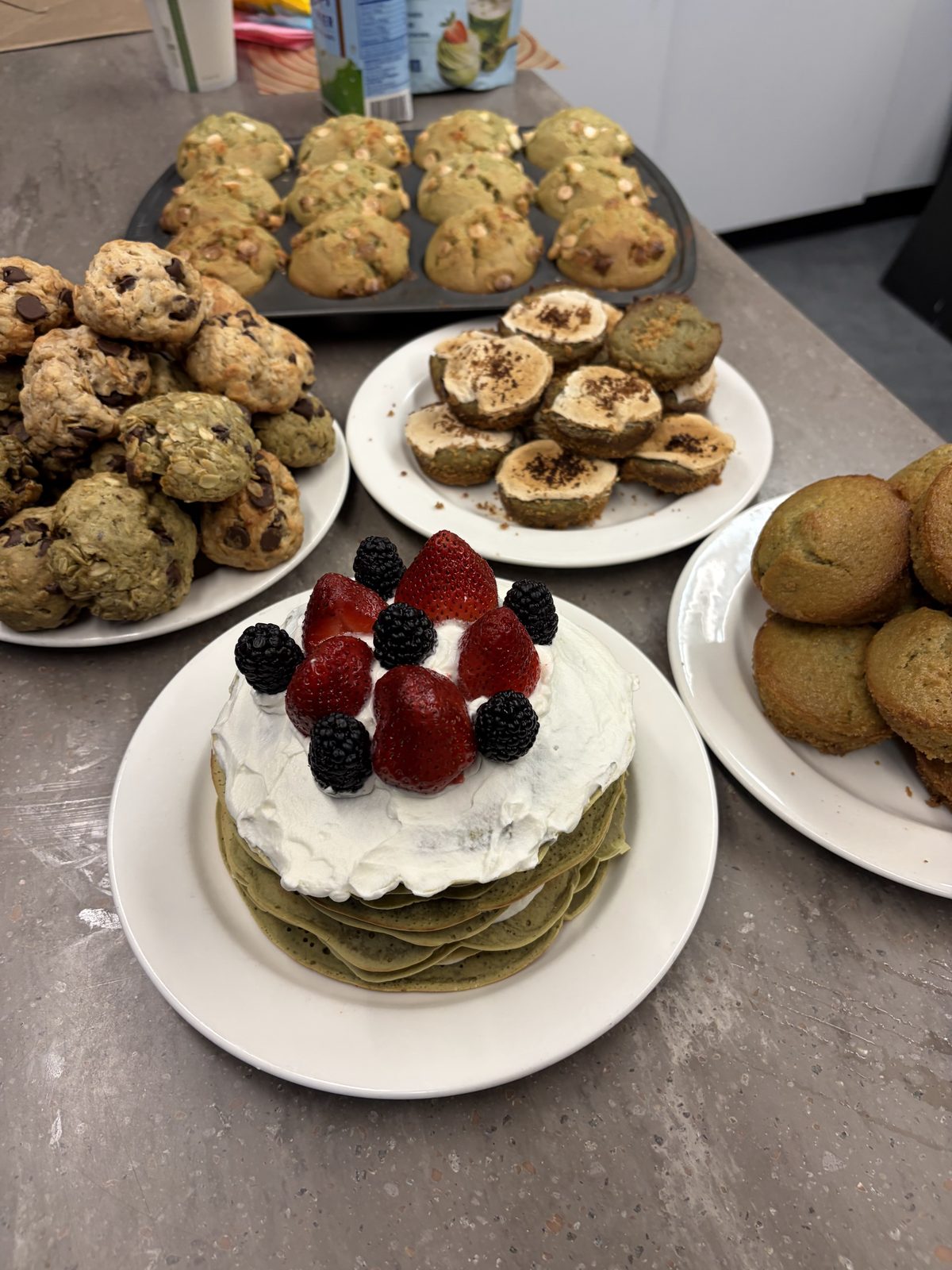 A table of baked goods: a green pancake stack topped with cream and berries, cookies, and muffins