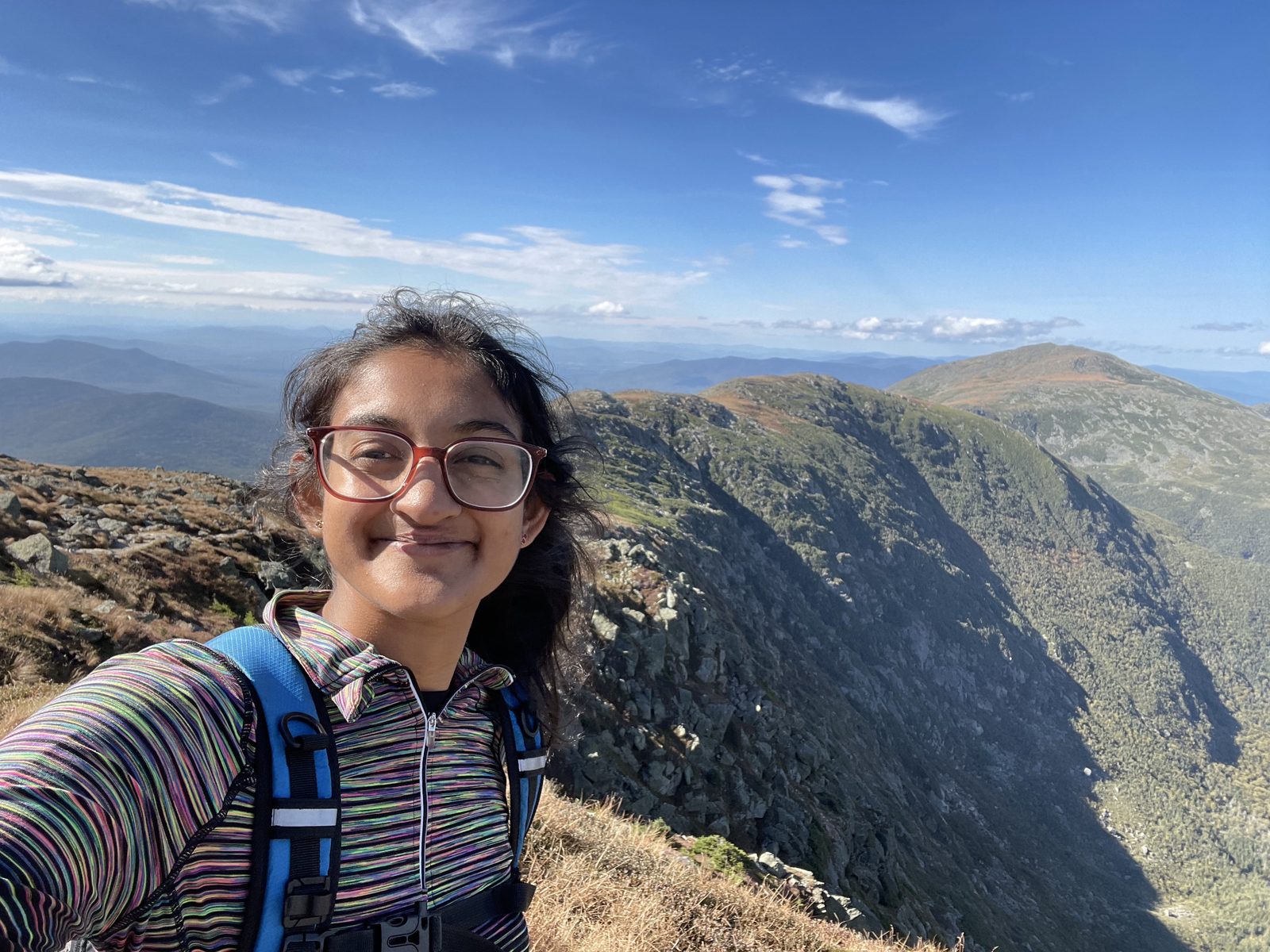 Suki smiling in a selfie on a rocky ridge above treeline, mountains behind